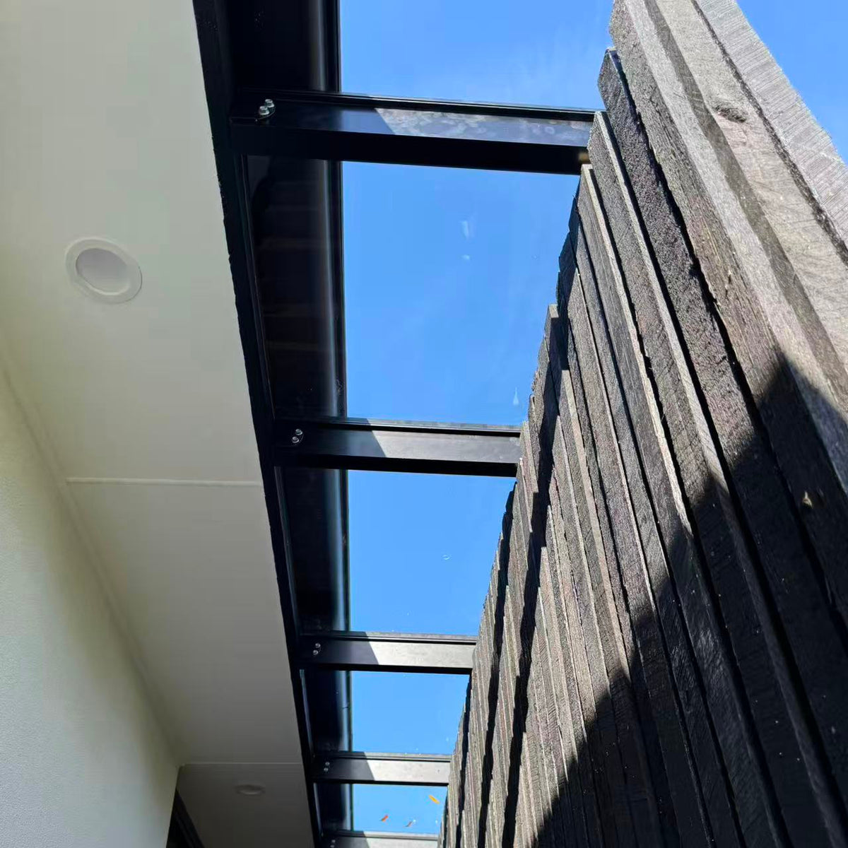 Modern outdoor patio pergola with a black metal frame and polycarbonate roof panels, supported by a dark wood slat wall under a clear blue sky.