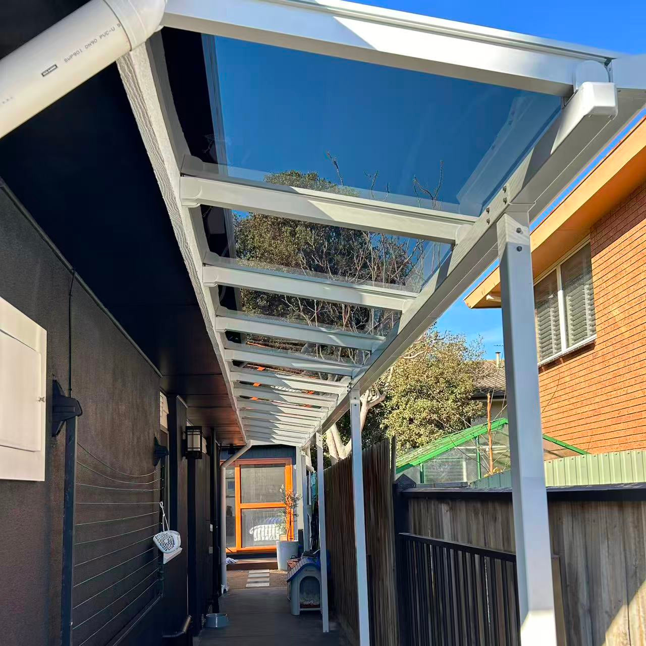 A white metal Aluminium pergola structure with a clear glass canopy installed over a narrow outdoor walkway between houses.