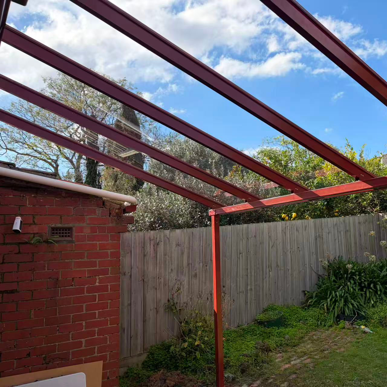A red metal Aluminium pergola kit installed in a backyard, with glass-like clear roofing sheets