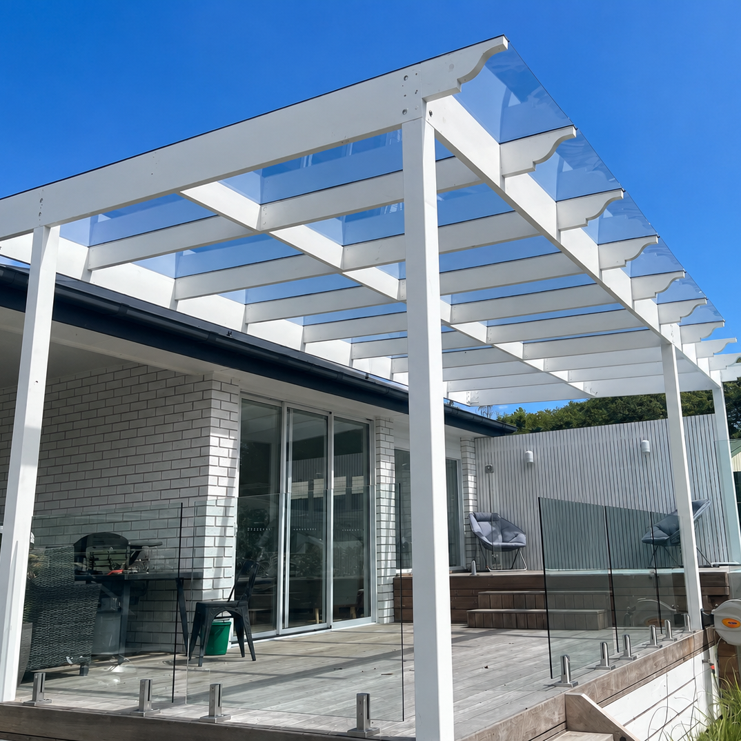 A modern white outdoor Skillion-roof pergola with clear glass panels installed over a wooden deck next to a house with glass sliding doors, under a bright blue sky.