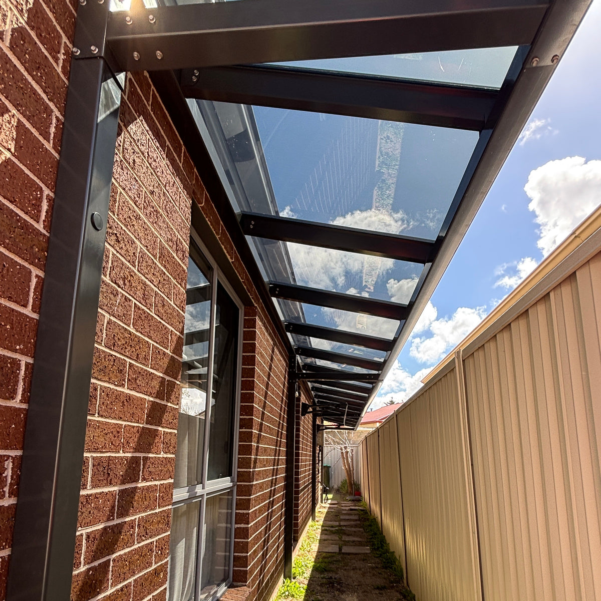 A modern outdoor walkway canopy with a clear glass roof and black metal frame, installed along the side of a red brick building next to a beige corrugated fence.
