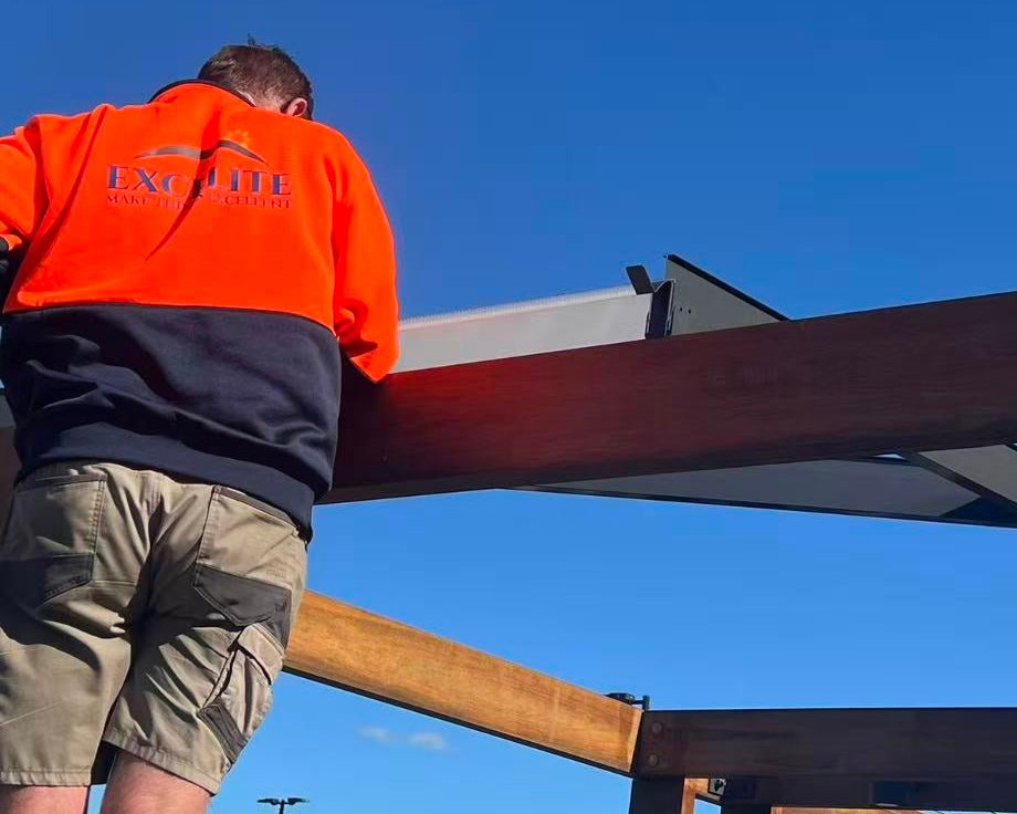 Excelite Staff wearing an orange and navy high-visibility work jacket and khaki shorts stands beside a large wooden pergola or roof structure