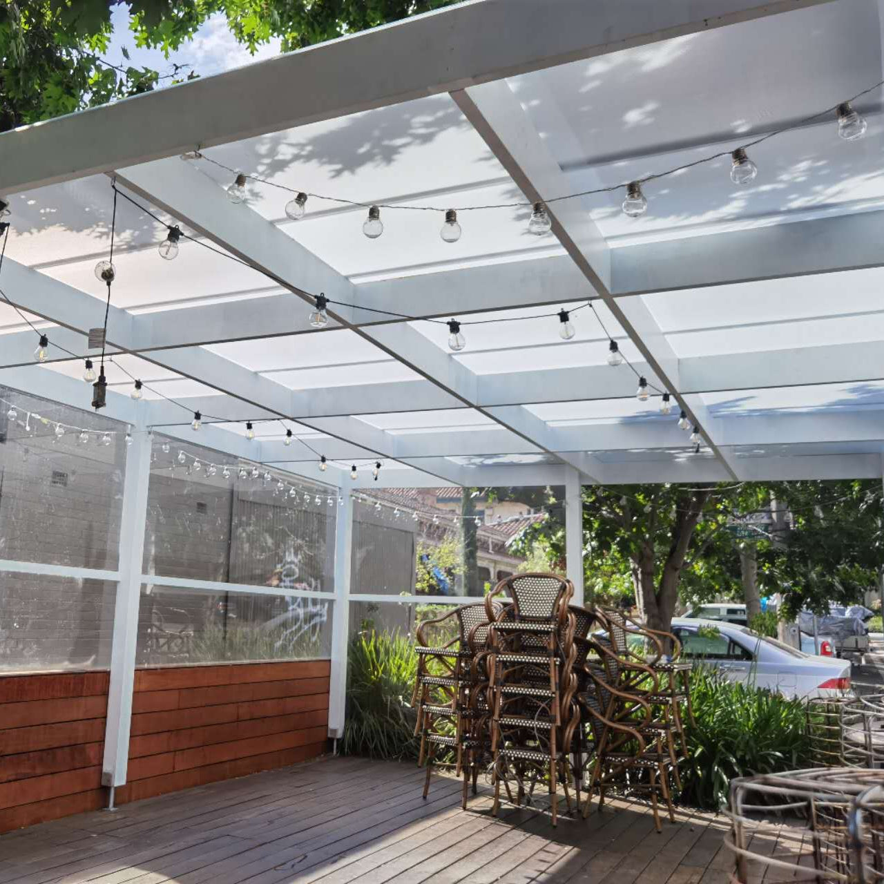 Outdoor patio pergola with a clear translucent roof, white support beams, string lights, a wooden deck, and stacked patio chairs near greenery.