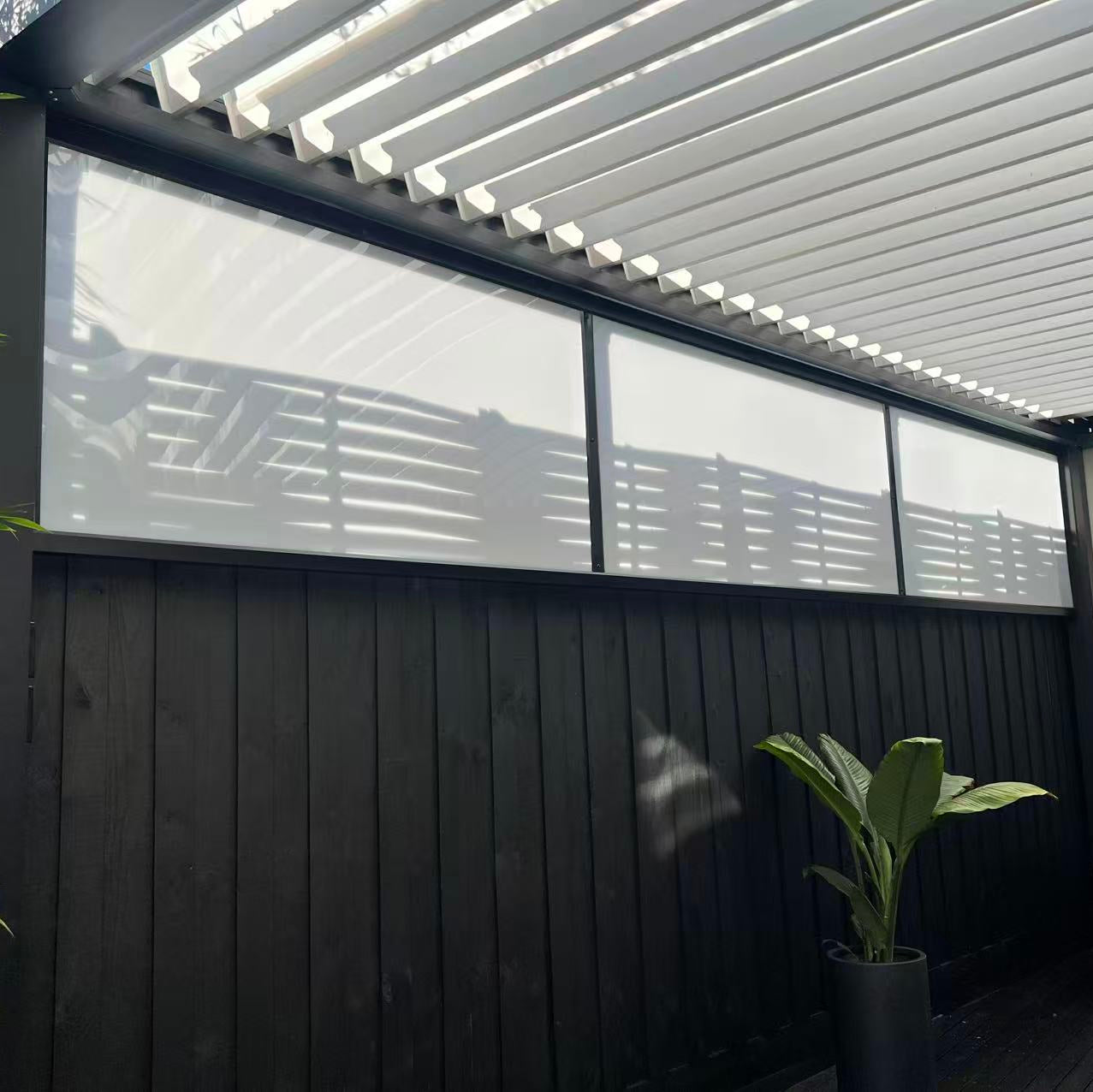 A modern outdoor patio area featuring a black wooden privacy fence with large frosted Translucent polycarbonate panels under a white slatted pergola ceiling. 