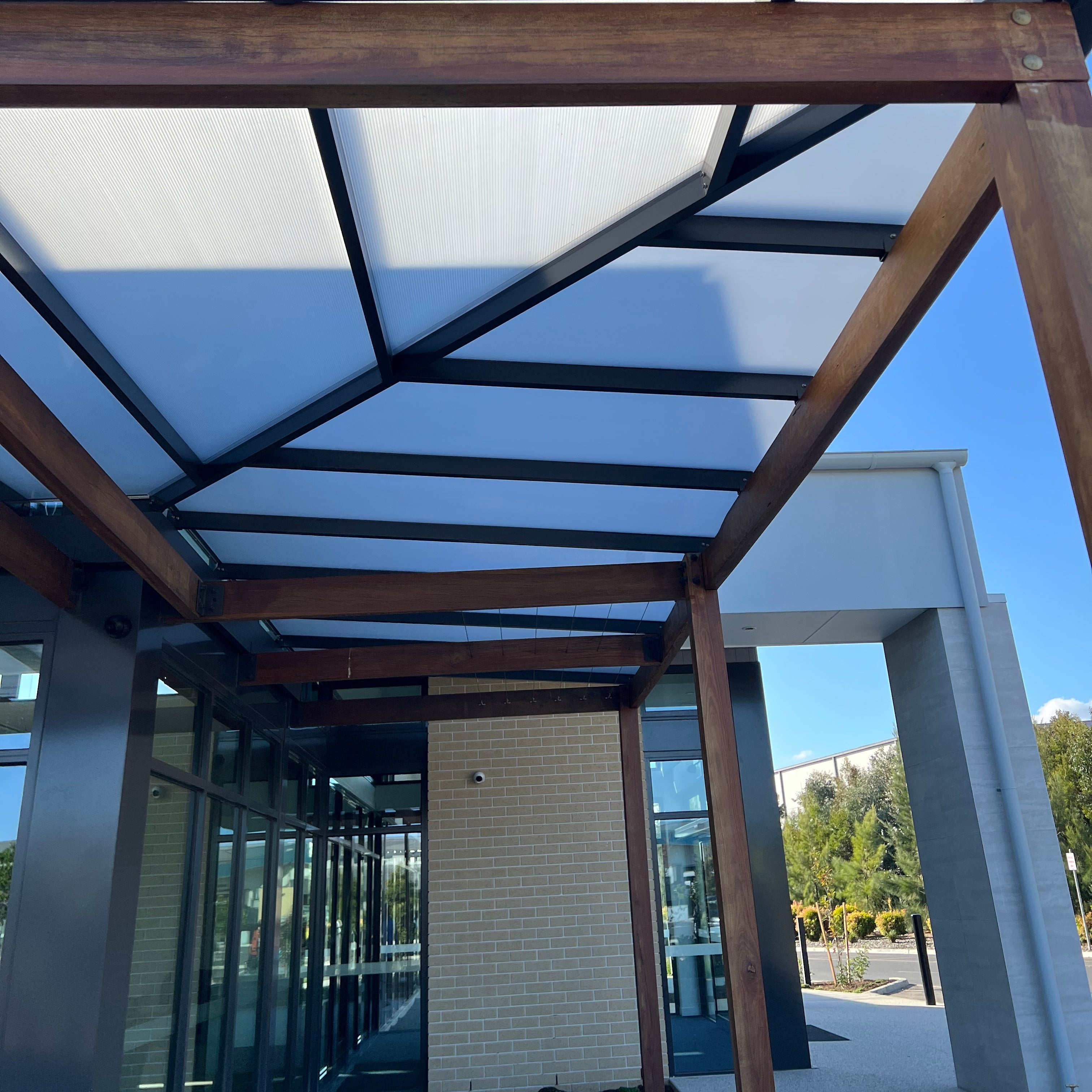 L shape Modern outdoor canopy structure with a geometric translucent roof panel system supported by dark metal beams and brown wooden posts.