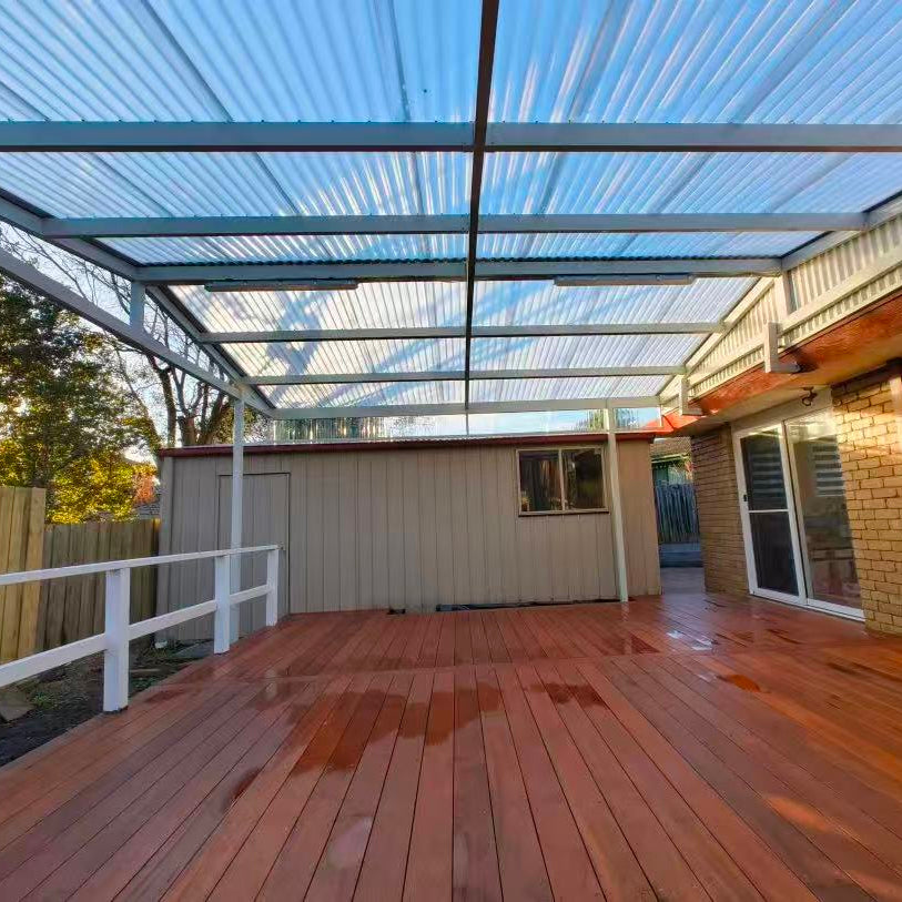 A large outdoor wooden deck with a Polycarbonate corrugated clear roof structure, shown next to a brick house with sliding glass doors and a fence on the left.